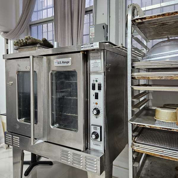 A stainless steel U.S. Range commercial oven and a metal rack with baking sheets in a dessert kitchen.