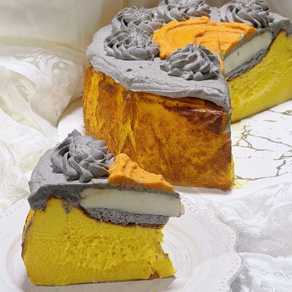A Studio Multisweet dessert with a slice removed, showing yellow cake, gray and white layers, and orange and gray frosting.