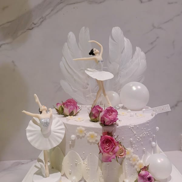 A Studio Multisweet dessert cake with two ballerina figurines, pink roses, white feathers, and pearls.