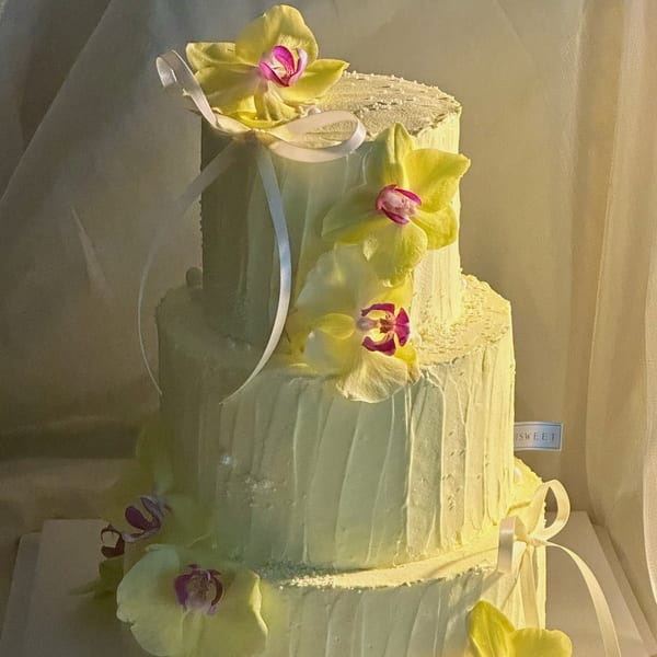 A three-tiered Studio Multisweet cake with pale yellow frosting, adorned with yellow orchids and white ribbons.