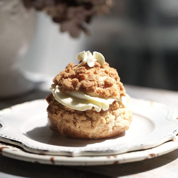 A Studio Multisweet cream puff with crumbly topping and white cream on a white plate.