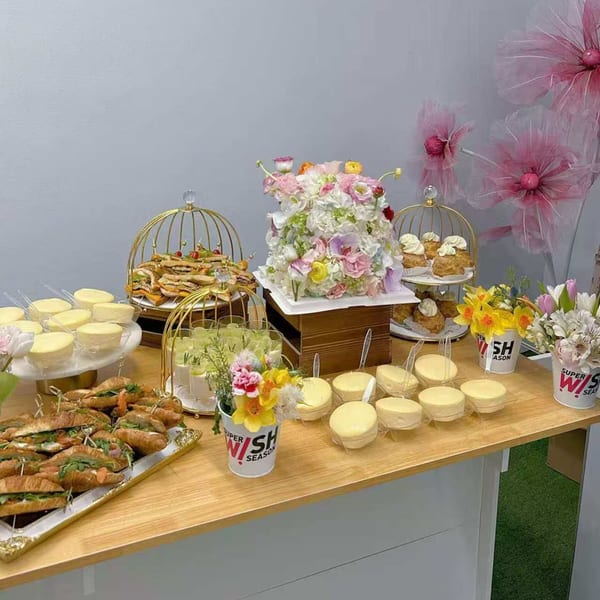 A dessert table with a floral cake, mini cheesecakes, sandwiches, and drinks, decorated with flowers.