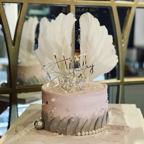 A Studio Multisweet birthday cake with pink and gray frosting, topped with white feathers, a crown, and "Happy Birthday" sign.