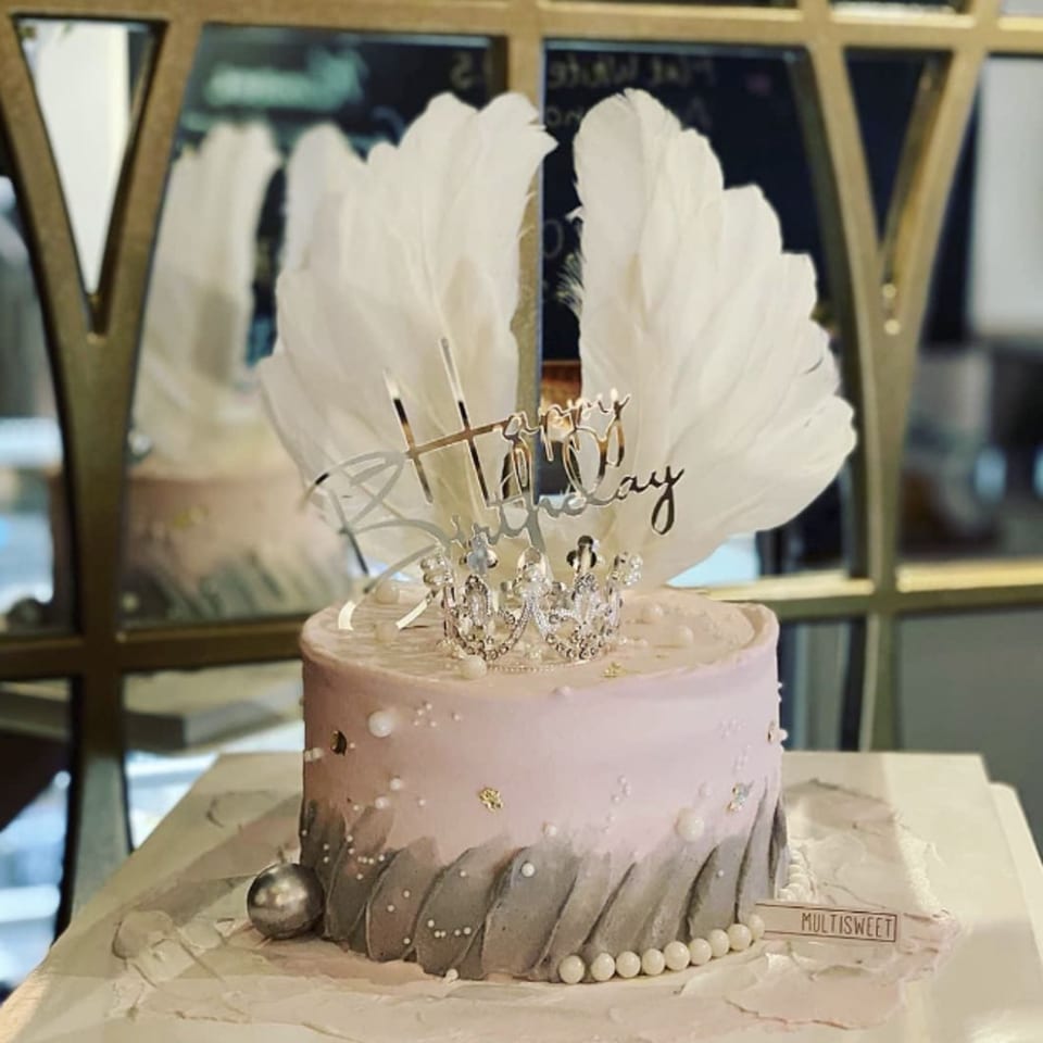 A Studio Multisweet birthday cake with pink and gray frosting, topped with white feathers, a crown, and "Happy Birthday" sign.