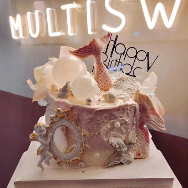 Studio Multisweet dessert cake with mermaid tail, angel, and "Happy Birthday" topper under neon sign.