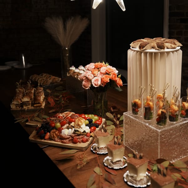 A dark, elegant buffet table laden with sliders, charcuterie, desserts, and floral arrangements.