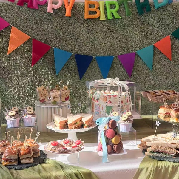 A birthday party spread with sandwiches, cupcakes, macarons, and a cake in a clear box.
