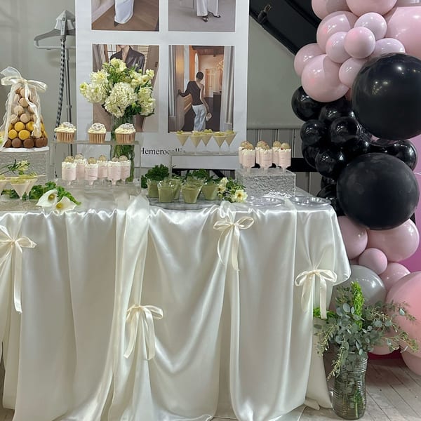 A dessert table with macarons, cupcakes, and various parfaits, decorated with white flowers and balloons.