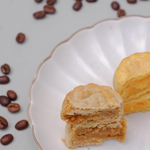 Two Studio Multisweet mooncakes, one cut open to reveal a golden filling, on a white plate with coffee beans.