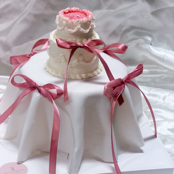 A two-tiered Studio Multisweet cake, white with pink frosting and ribbons, on a white draped surface.