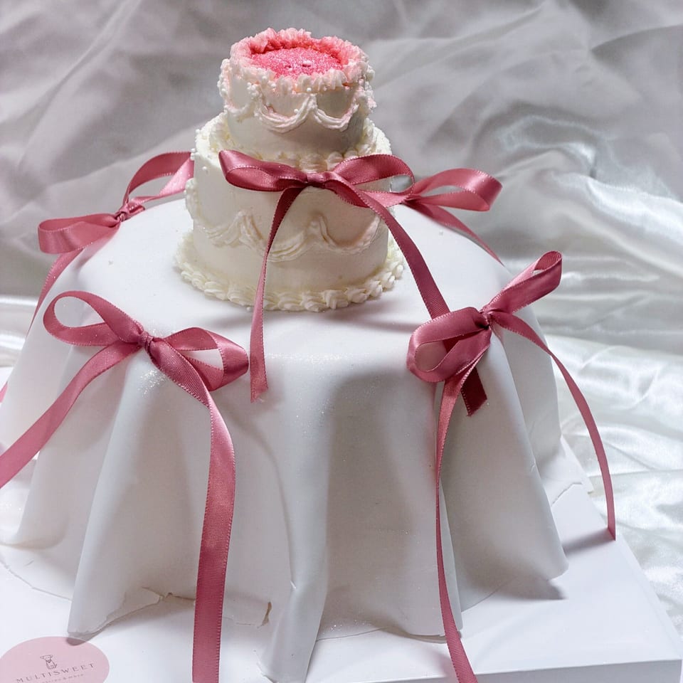 A two-tiered Studio Multisweet cake, white with pink frosting and ribbons, on a white draped surface.