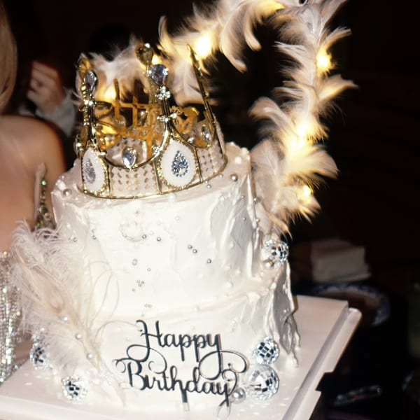A two-tier white birthday cake with a gold crown, feathers, disco balls, and "Happy Birthday" topper.