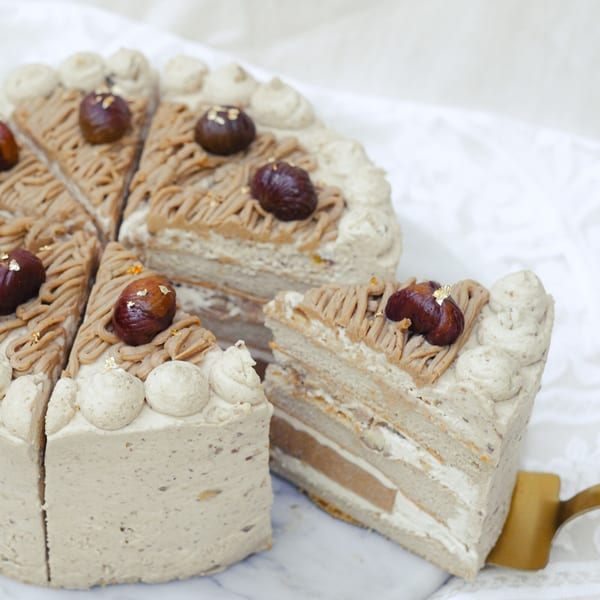 A sliced chestnut cake with cream frosting and whole chestnuts on top, with a slice being served.