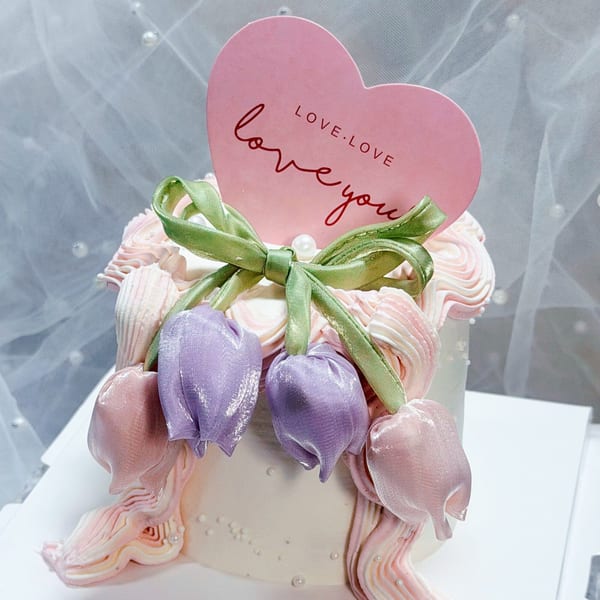 White cake with pink and purple fabric tulips, a green ribbon, and a pink heart-shaped "Love You" topper.