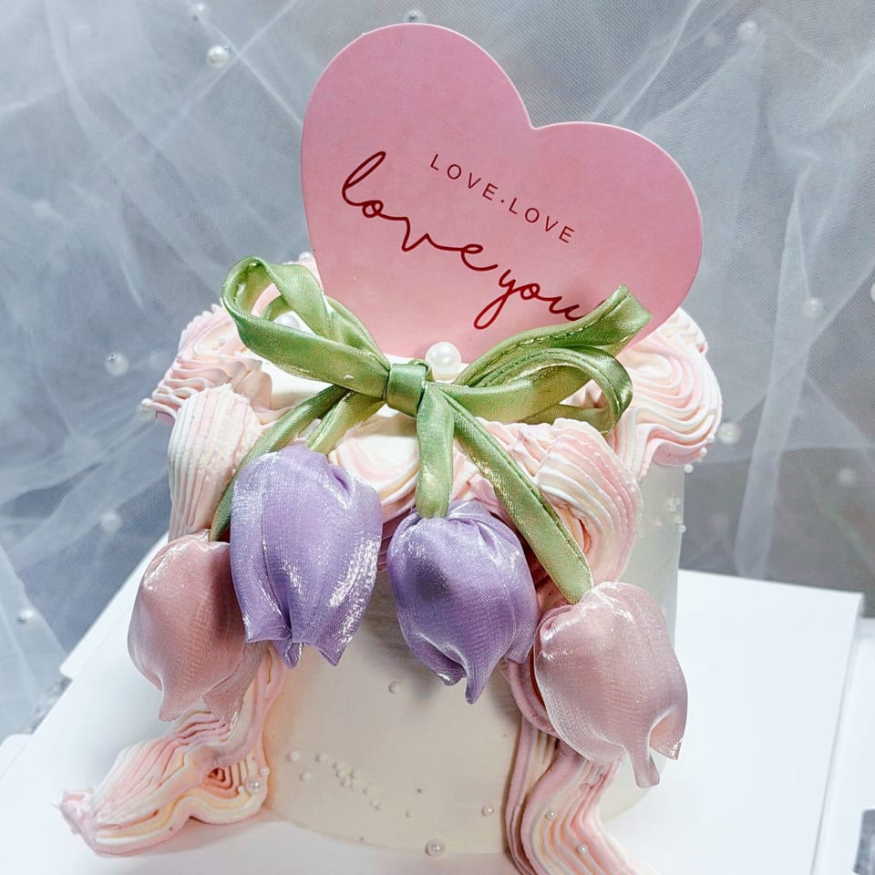 White cake with pink and purple fabric tulips, a green ribbon, and a pink heart-shaped "Love You" topper.