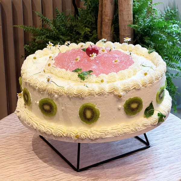 A large white cake with kiwi slices, pink jelly, and small white flowers on a stand.