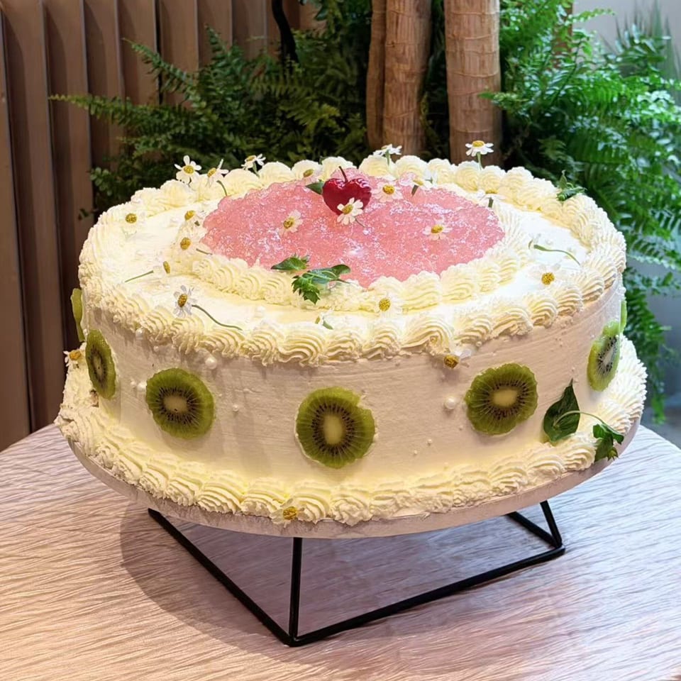 A large white cake with kiwi slices, pink jelly, and small white flowers on a stand.