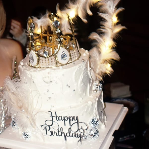 A two-tiered white birthday cake with a golden crown, feathers, and disco ball decorations.
