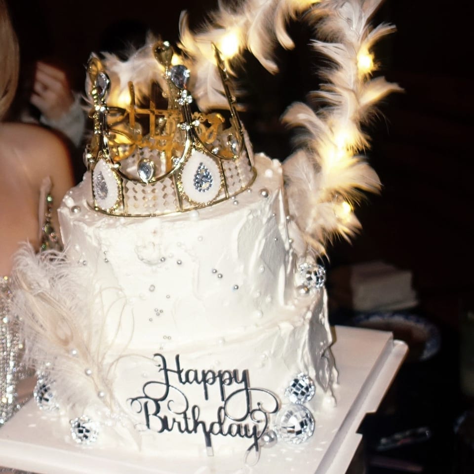 A two-tiered white birthday cake with a golden crown, feathers, and disco ball decorations.
