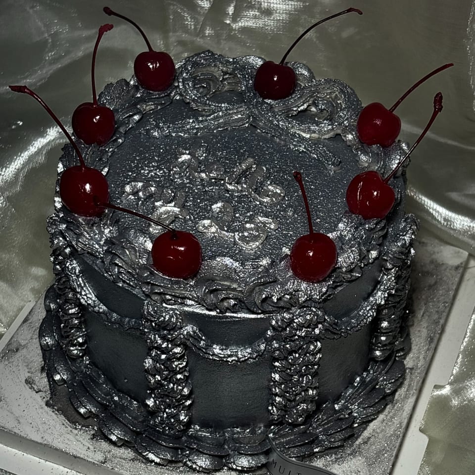 A Studio Multisweet dessert, a round silver cake with ornate frosting and eight red cherries on top.