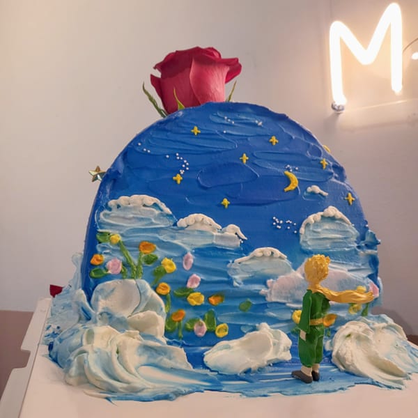 A blue, dome-shaped cake with a red rose on top, decorated with stars, clouds, and a figurine.